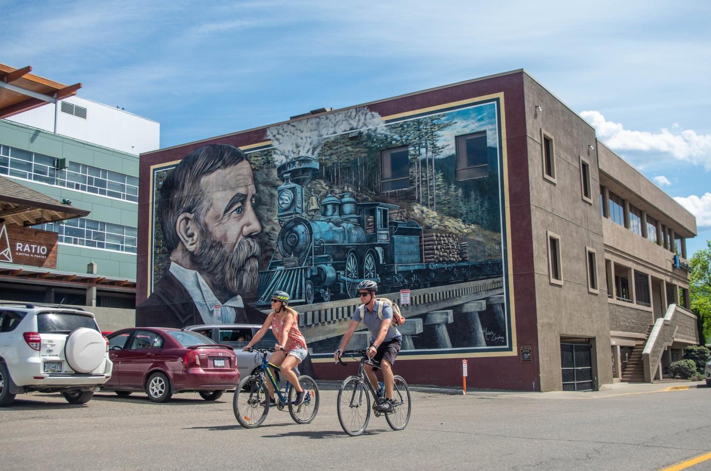 Man and woman biking downtown Vernon passed a painted mural on a building of a train and train tracks
