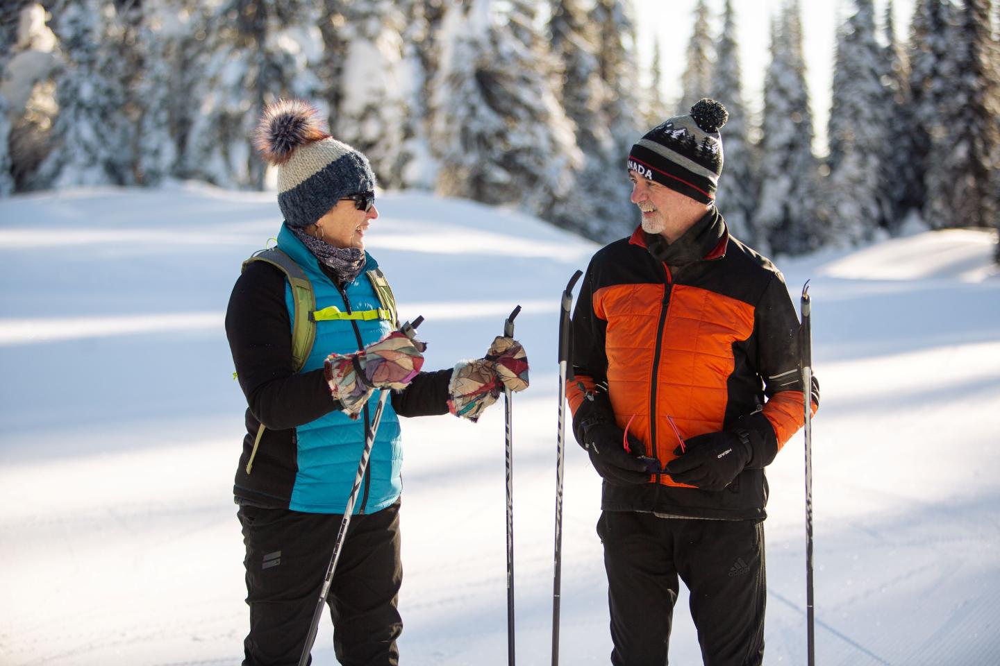 Two people cross-country skiing in a snowy forest, smiling and talking.