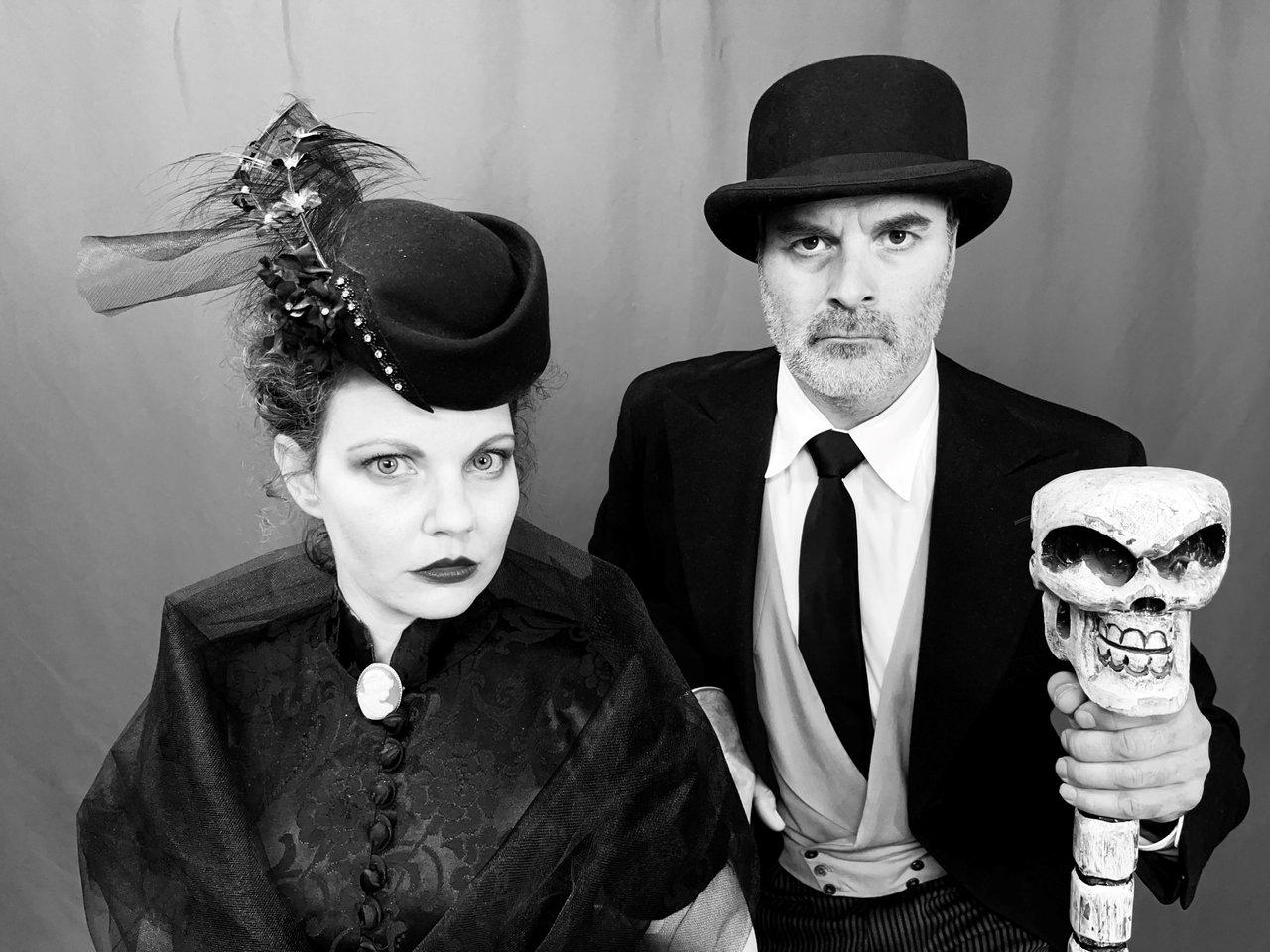Man and woman in vintage attire, woman in hat, man with top hat and skull-topped cane, black and white.
