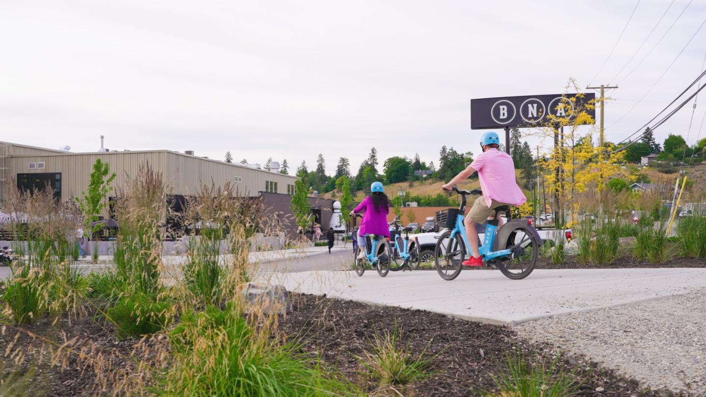 Two people on bikes ride along a path, surrounded by greenery and buildings.