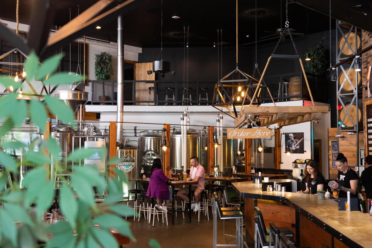 Modern café interior with people sitting, warm lighting, and green plant in foreground.