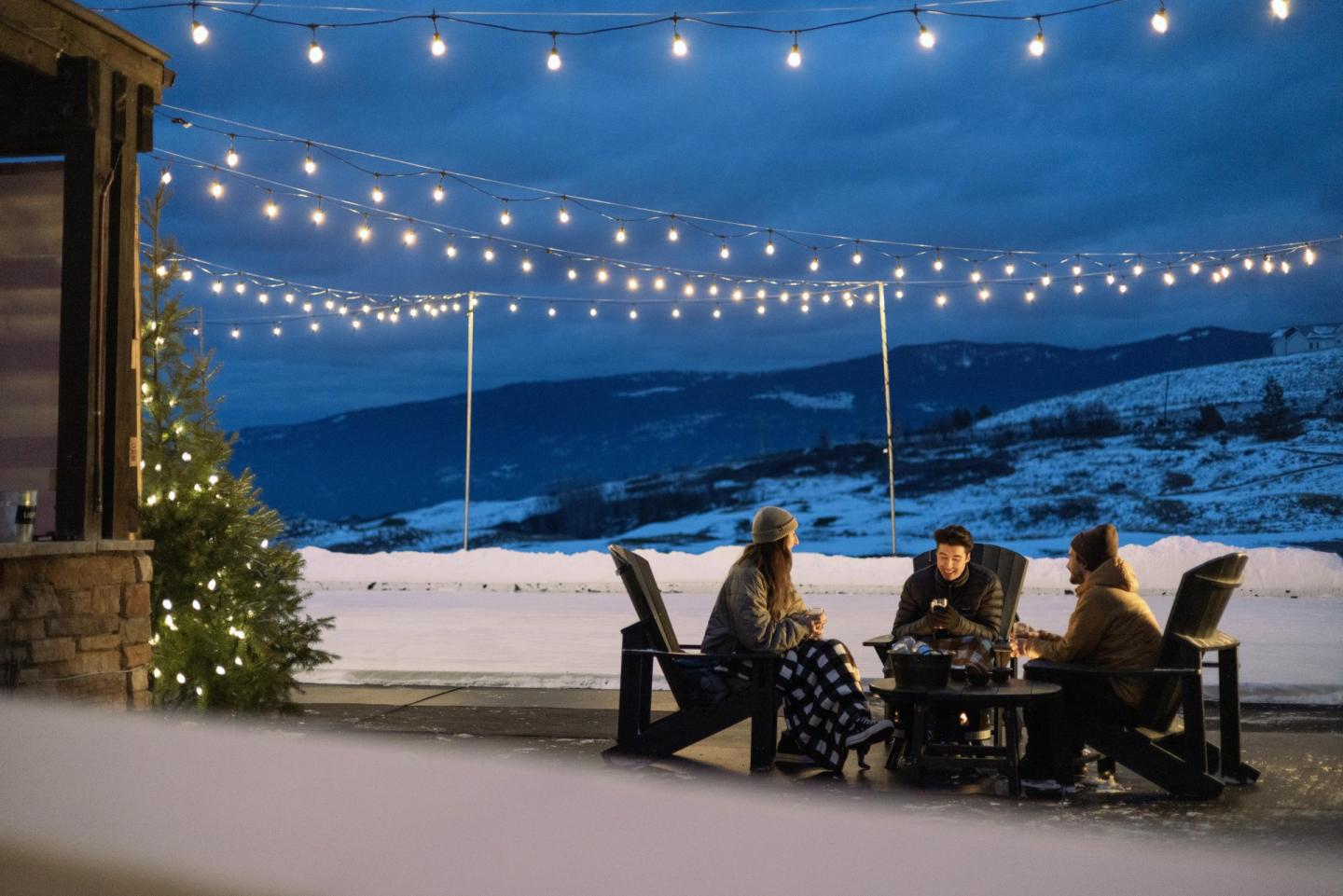 Friends gather around a fire pit under string lights in a snowy landscape.