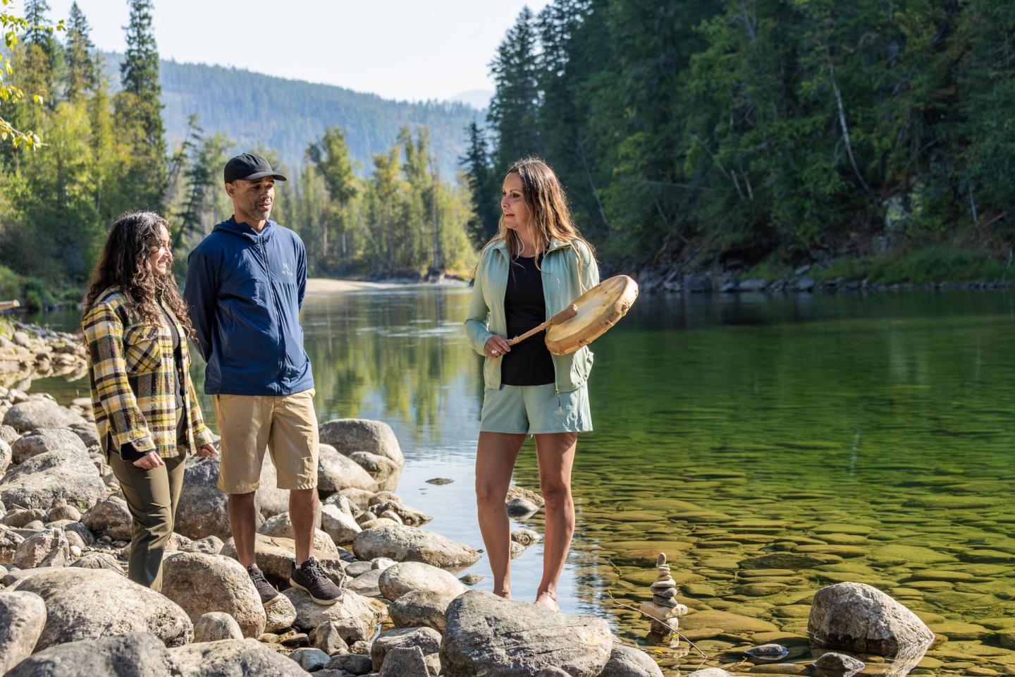 Three people stand on a rocky riverbank, with trees in the background.