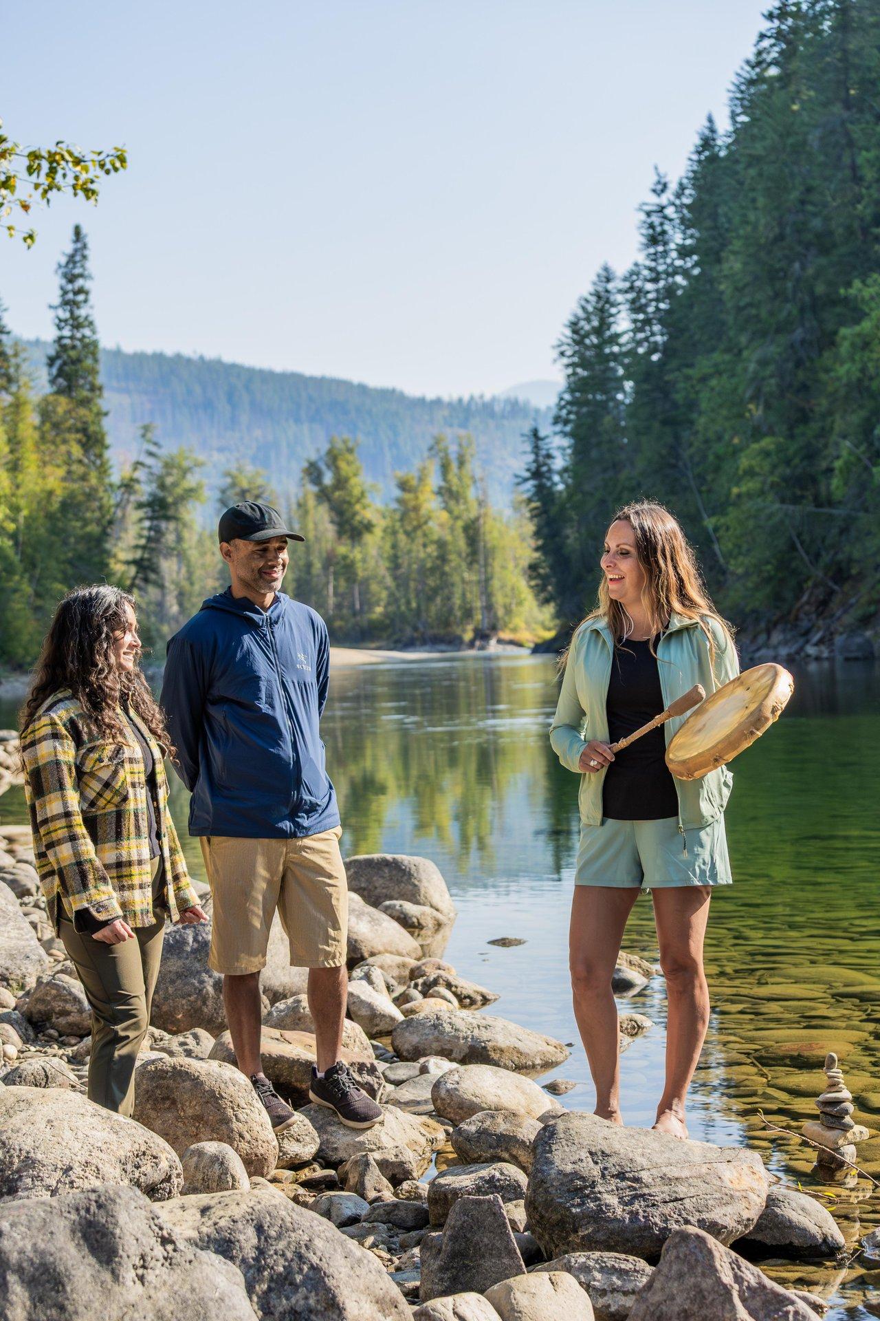 Three people talking by a river, surrounded by trees and rocks.