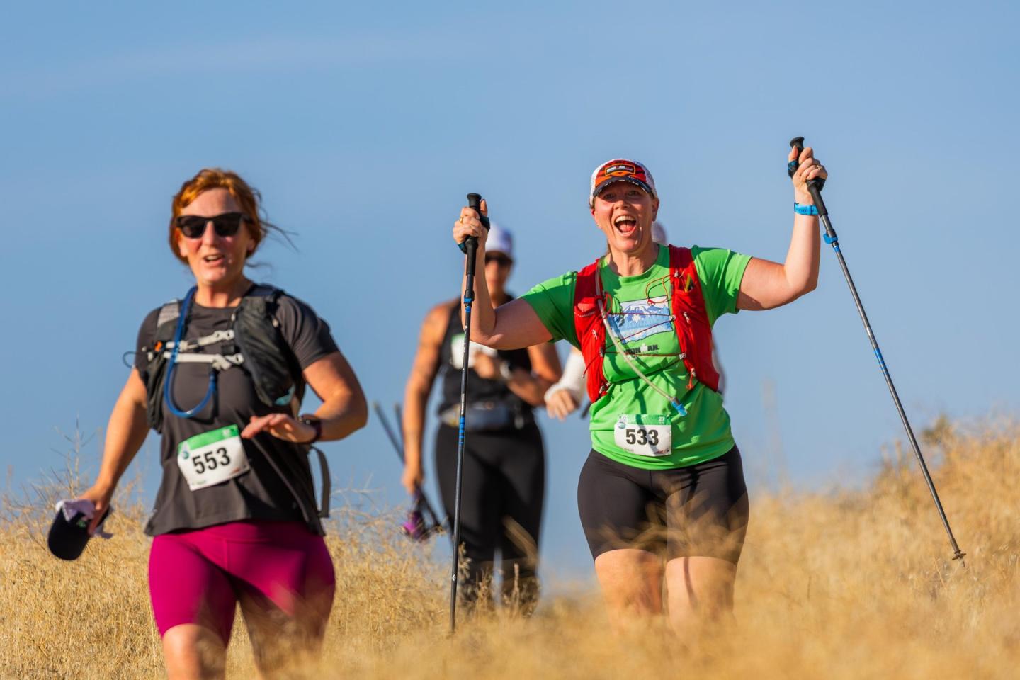 Trail runners with poles in a dry, grassy landscape under a clear blue sky.